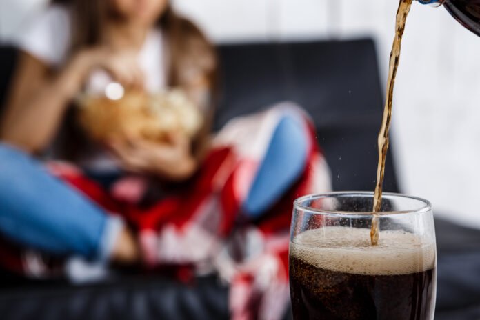 Girl eating chips, sitting on sofa. Fosuc  glass of soda. Mais de 100 mil novos casos de Diabetes seriam evitados se fosse eliminado o consumo de bebidas como: refrigerantes, sucos diet, light.